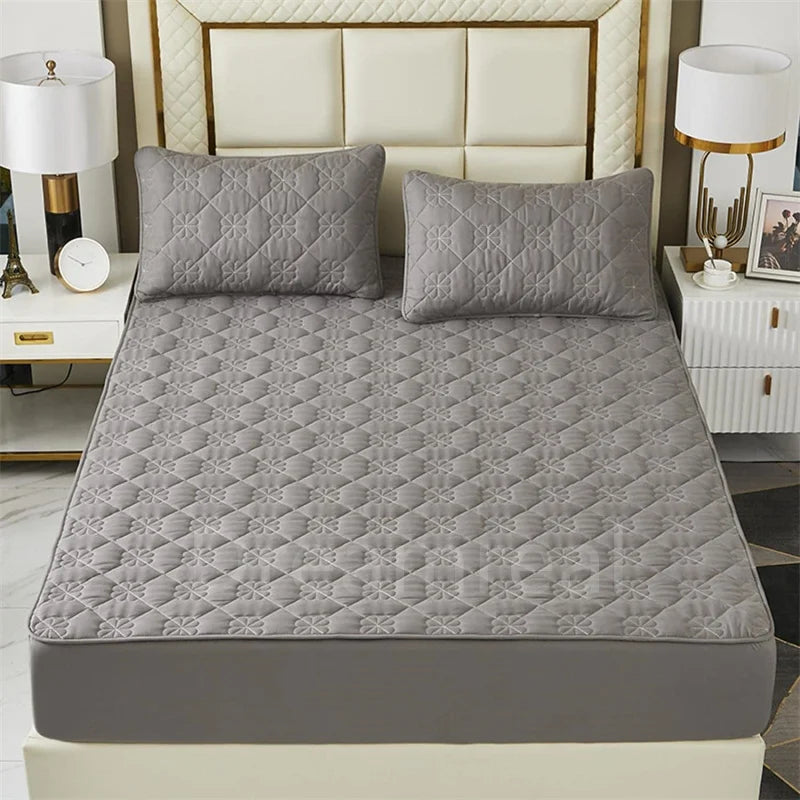 A gray quilted mattress protector displayed on a bed with a light upholstered headboard and bedside tables.