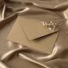 A beige linen texture envelope is placed on draped satin fabric with a small sprig of white flowers.