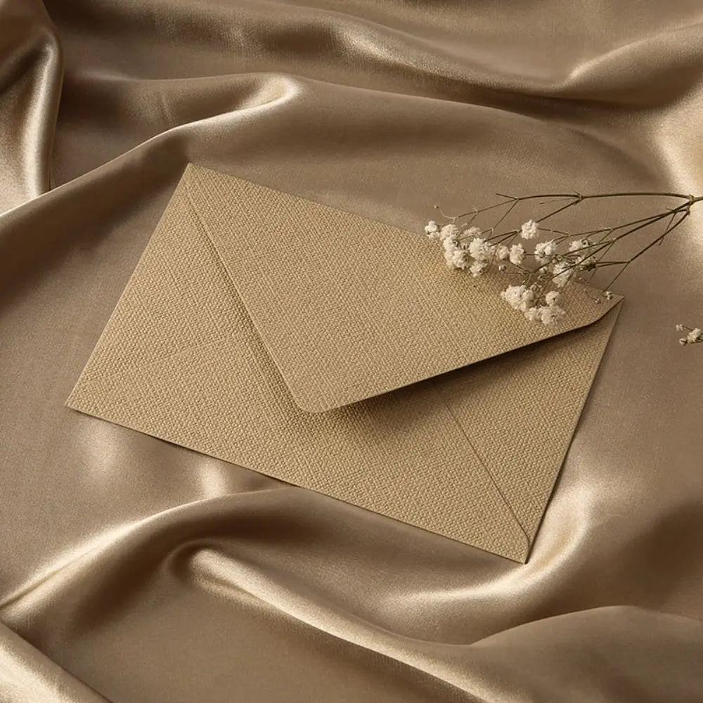 A beige linen texture envelope is placed on draped satin fabric with a small sprig of white flowers.