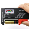 A hand holds a black battery tester device with a red button and a gold battery cell inserted.
