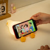 A hand holds a smartphone displaying a cartoon, with silicone duck shapes nearby.