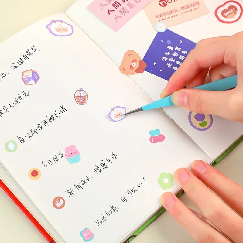 A hand uses a fine-tipped tool to apply colored glue or paint onto a page of a scrapbook or journal featuring small macaron i