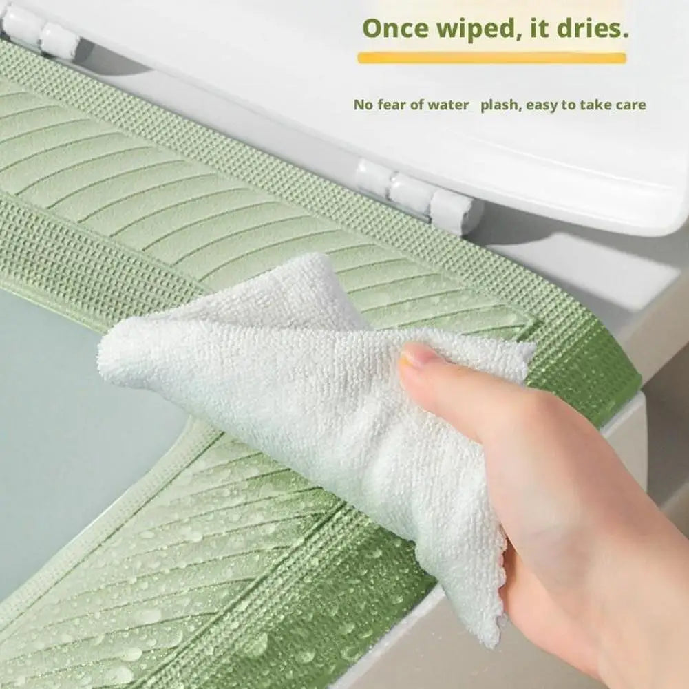 A hand is wiping a white, textured cleaning cloth on a light green toilet seat cover.
