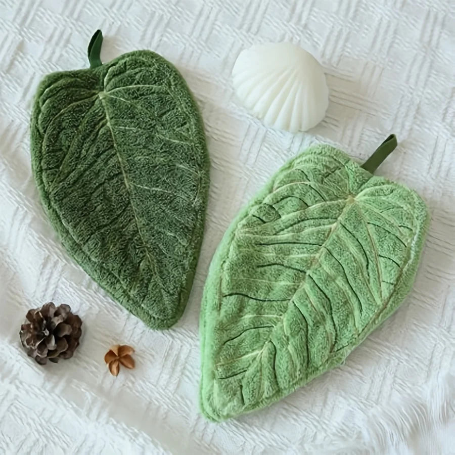 A green leaf-shaped coral fleece hand towel is displayed on a white surface next to a seashell and a pinecone.