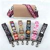 A beige crossbody bag with a purple and pink floral patterned strap is shown alongside several other colorful patterned strap