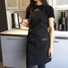 A woman wearing a black durable canvas kitchen apron is standing in a kitchen preparing to use a silver coffee grinder.