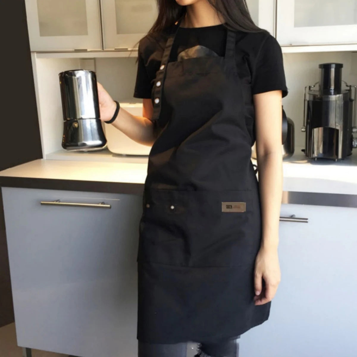 A woman wearing a black durable canvas kitchen apron is standing in a kitchen preparing to use a silver coffee grinder.