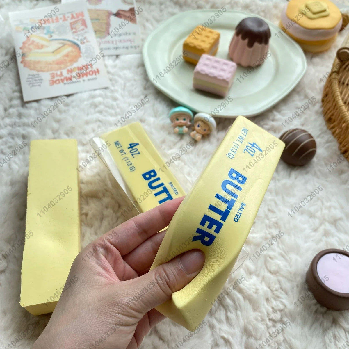 Eine Hand hält ein gelbes Butterriegel-Squeeze-Fidget-Spielzeug neben verschiedenen butterthematischen Leckereien und Süßigke