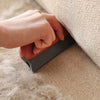 A hand is using a gray pet hair removal brush on a beige carpet, demonstrating its use for grooming pets.