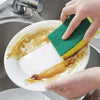 A hand is using a green and yellow double-sided cleaning sponge to wash food residue from a white plate in a kitchen sink.