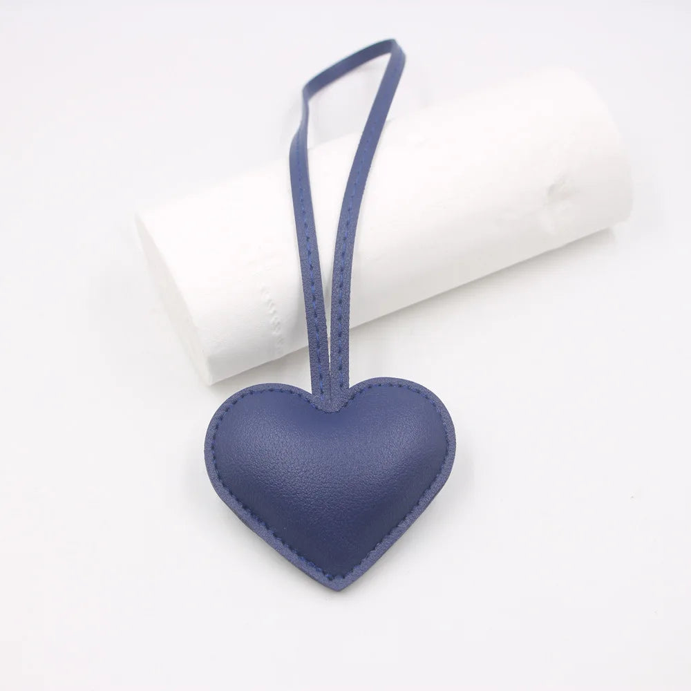A dark blue heart-shaped PU leather keychain with a thin strap and a small cotton charm, resting on a white cylindrical objec