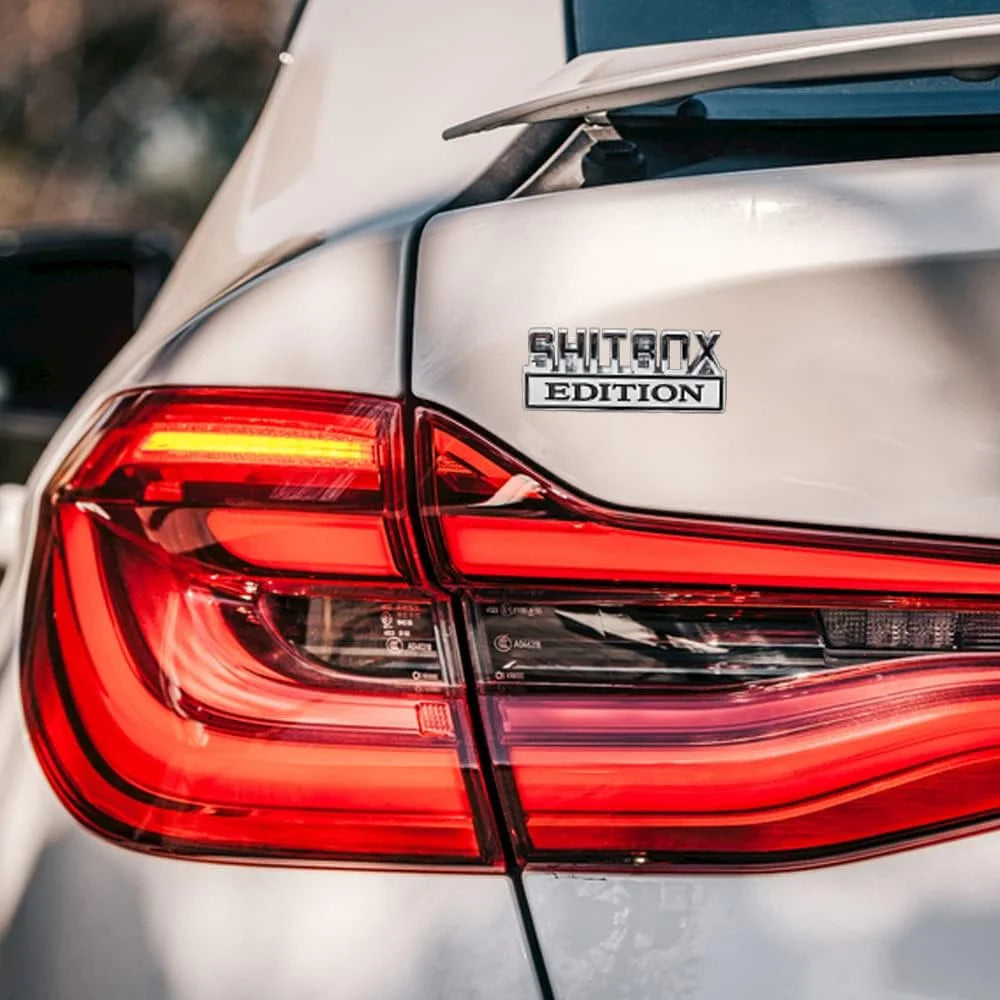 Nahaufnahme der Rückleuchte eines silbernen Autos mit einem SHITBOX EDITION Aufkleber-Badge.