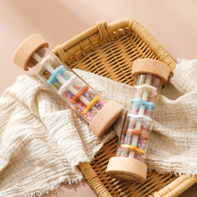 A wooden rain rattle sensory toy in glass bottles with colorful beads resting on a woven tray and linen fabric.