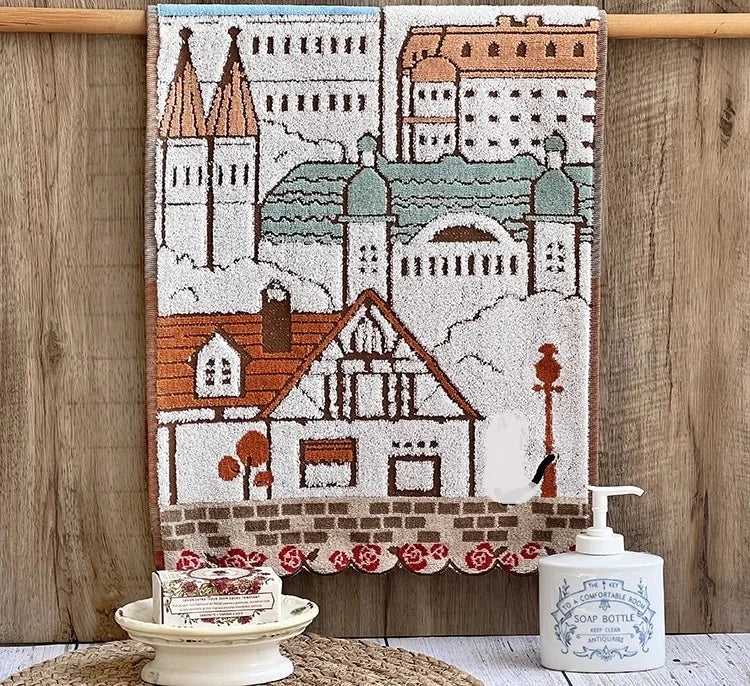 A rectangular embroidered cartoon face towel featuring a cityscape and a house, hanging on a wooden rod.