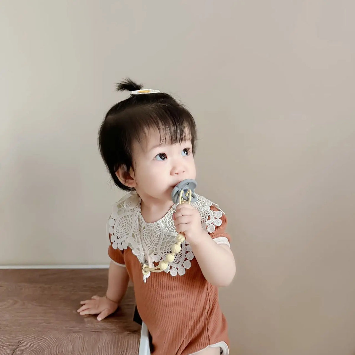 A baby is holding a silicone pacifier clip with a teether, looking to the side.