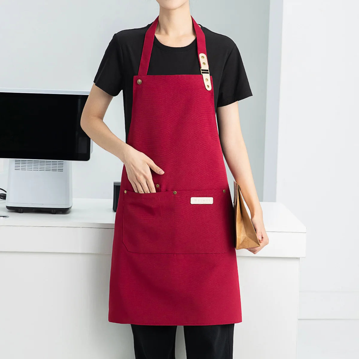 A woman wearing a deep red waterproof canvas chef apron over a black shirt, standing in a kitchen setting.