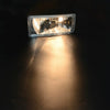 A clear automotive fog light assembly is illuminated, casting a bright beam onto a dark surface.