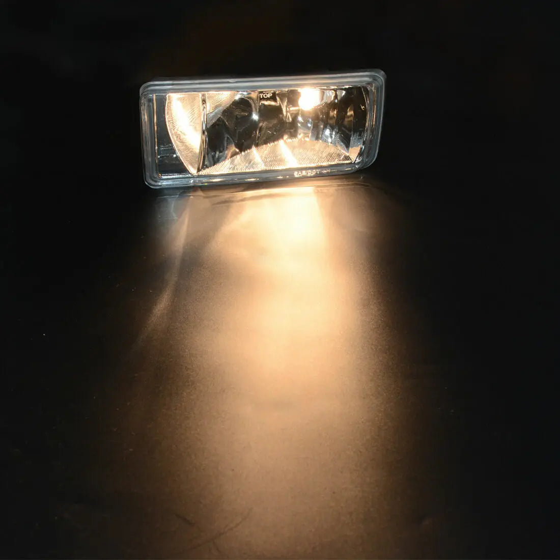 A clear automotive fog light assembly is illuminated, casting a bright beam onto a dark surface.