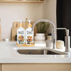 A glass bottle of hands soap and a bottle of dishes soap with pump dispensers resting on a wooden tray above a kitchen sink.