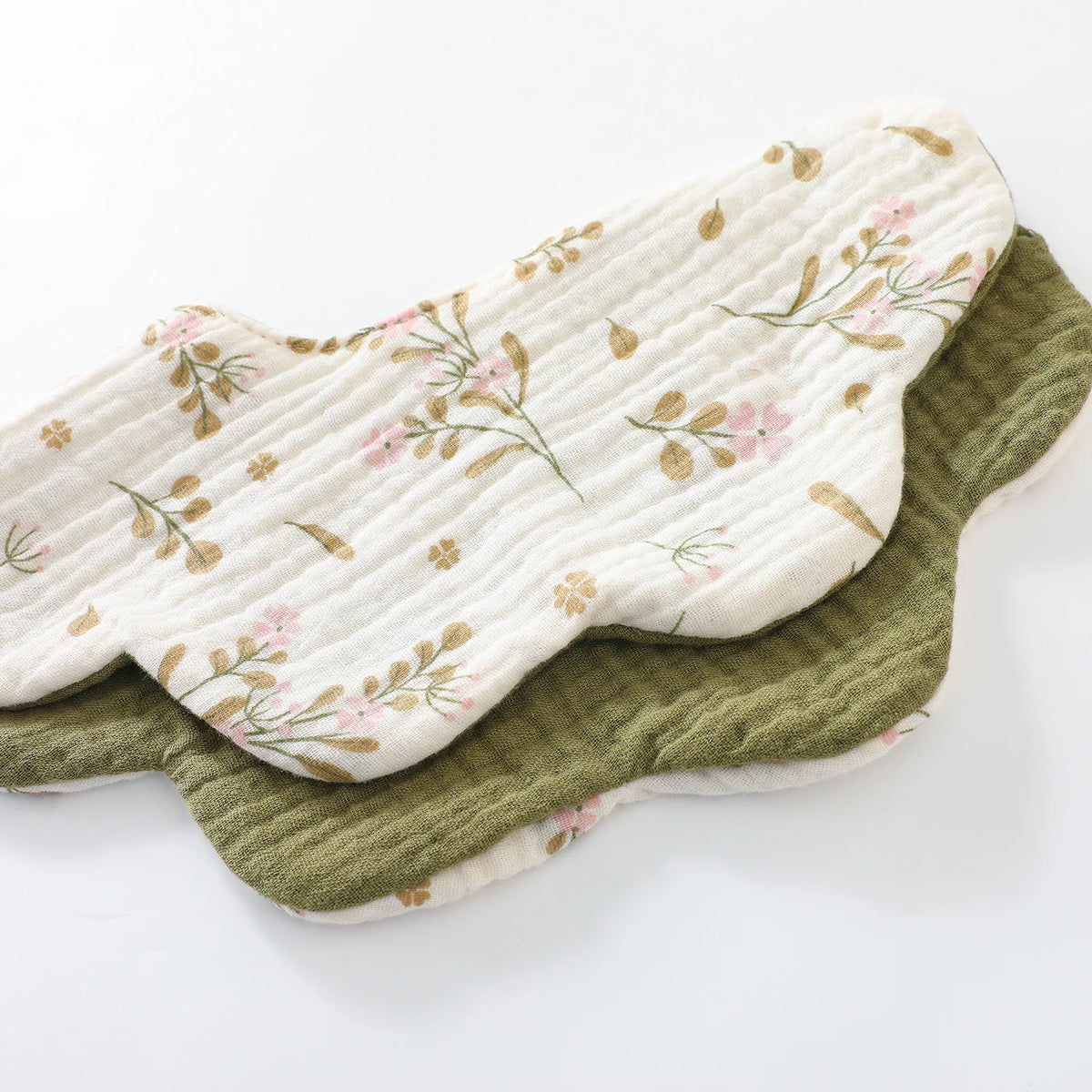 A light cream cotton baby feeding bib with a floral pattern and a darker olive green absorbent towel stacked together.