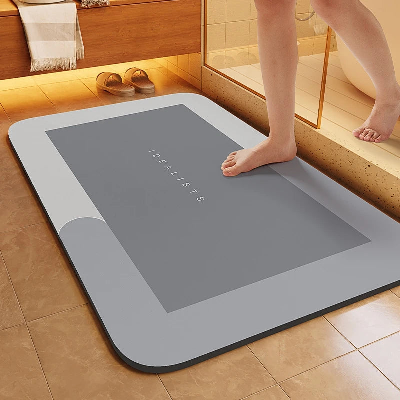 A gray non-slip bath mat with the word IDEALISTS printed on it is placed on a tiled floor next to a person's bare feet.