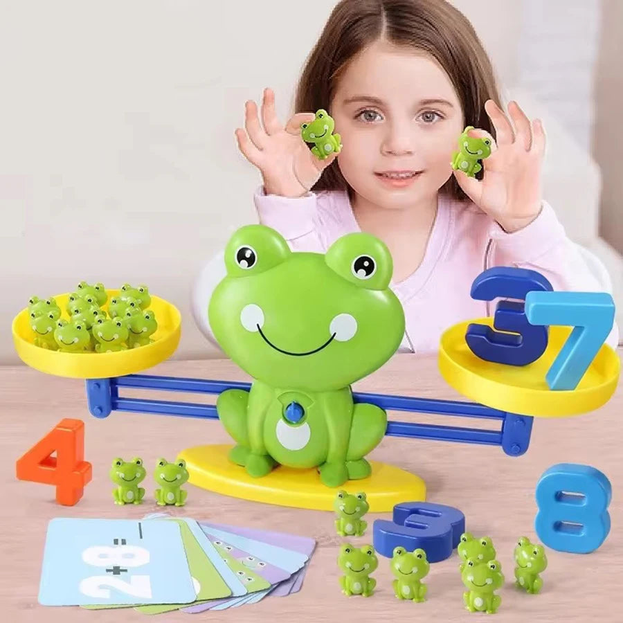 A young girl is holding a green frog toy while surrounded by yellow and blue balance math game components.