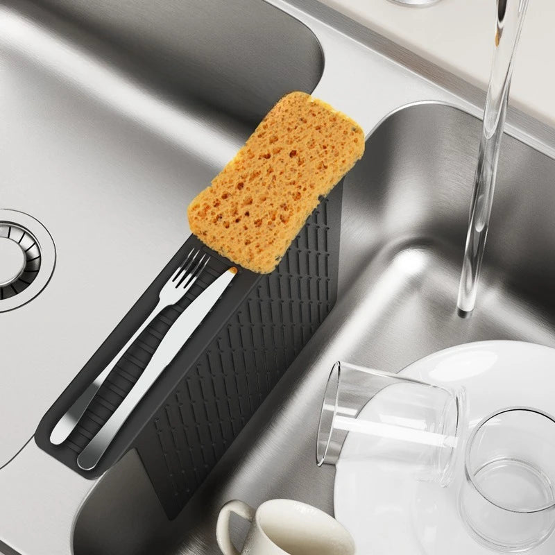 A silicone sink protector mat with drainage holes is shown in a stainless steel sink, featuring a fork and a piece of orange-