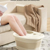 A beige foldable foot bath tub is shown being used by a person on a sofa, with a towel draped nearby.
