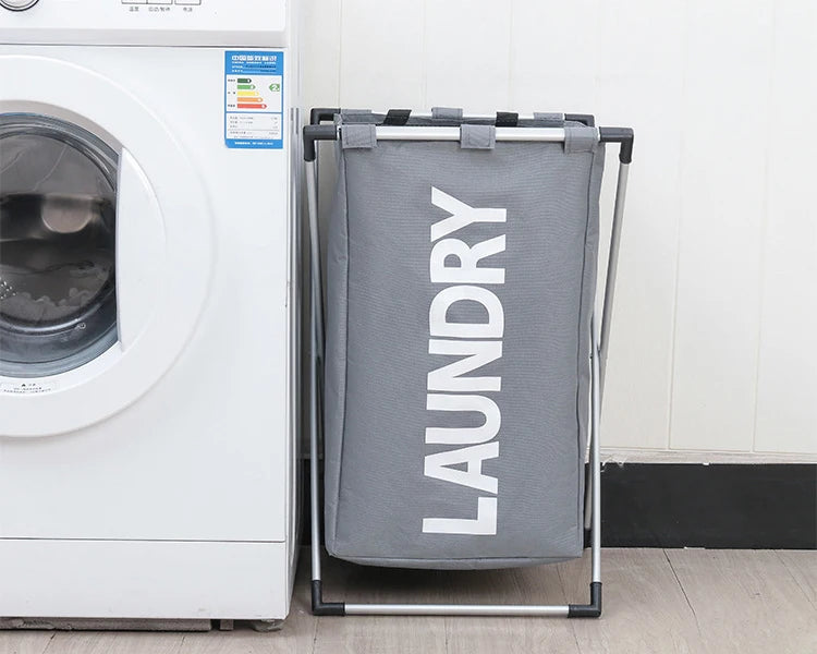 A gray foldable laundry basket with a metal frame is shown next to a white washing machine.