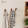Three walnut wood hooks are mounted on a wall, displaying a gray towel, a black and white patterned scarf, and a light gray h