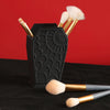 Black coffin-shaped makeup brush holder with a spider web design and several gold-handled makeup brushes.