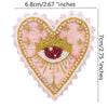 A heart-shaped patch embroidered with gold and pearl beads featuring a stylized eye design on pink fabric.