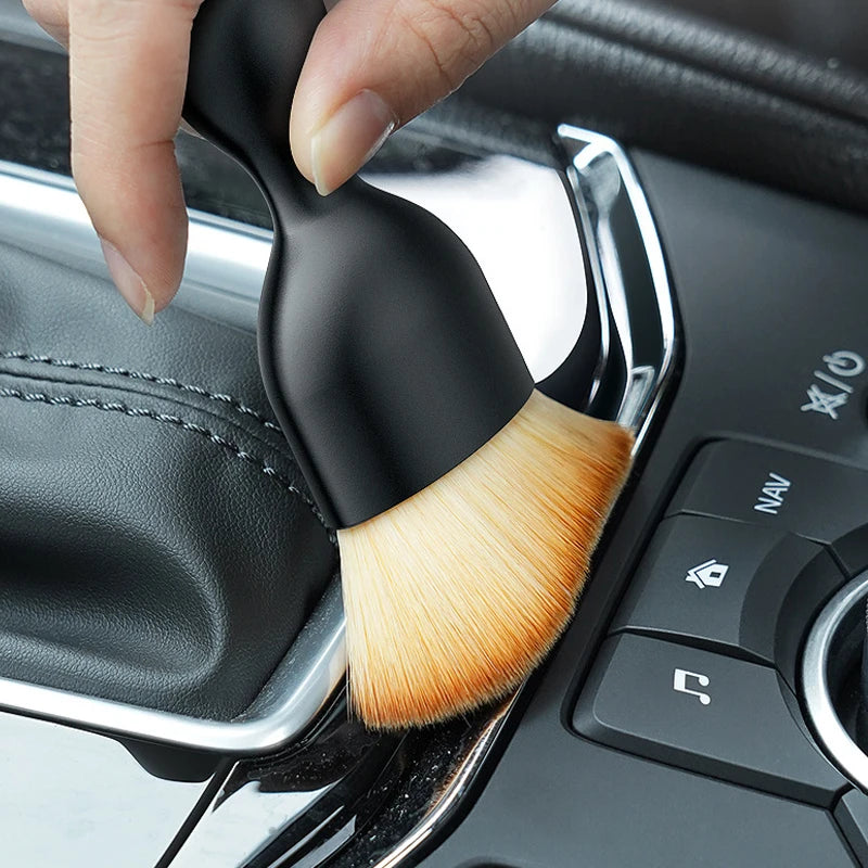 A hand is holding a black soft-bristle cleaning brush with a protective case, positioned near a car vent or crevice.