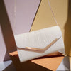A white pleated glitter envelope clutch with a gold-toned chain strap resting on an orange surface.