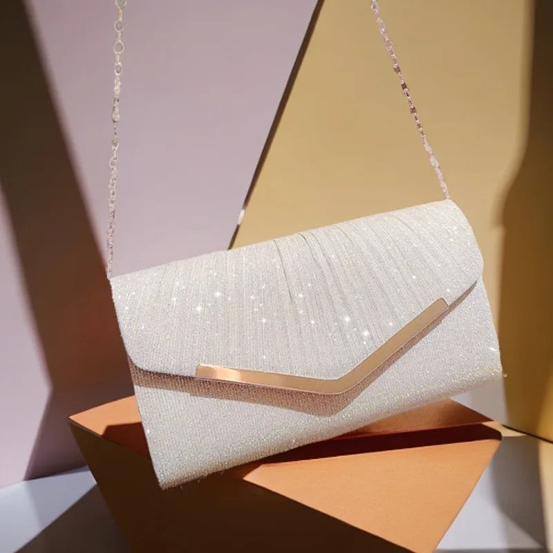A white pleated glitter envelope clutch with a gold-toned chain strap resting on an orange surface.