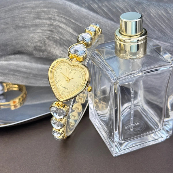 Goldfarbener, herzförmiger Quarz-Armbanduhrarmreif neben einer klaren Glas-Parfümflasche.