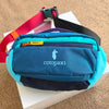 A teal crossbody chest bag with a black strap and multiple compartments resting on a white box.