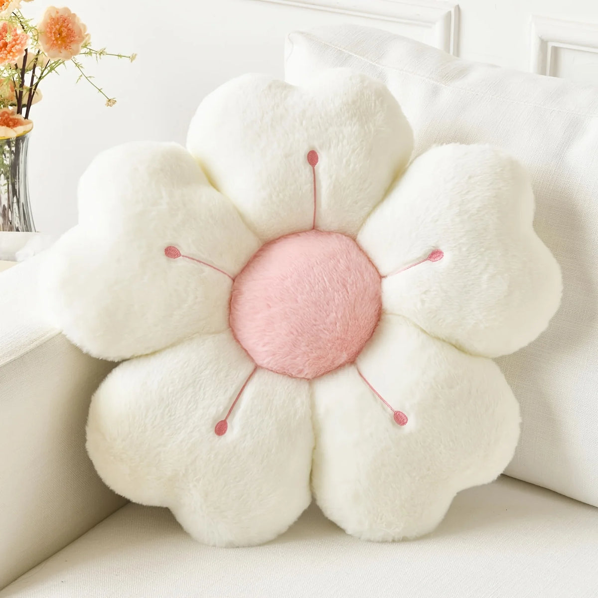 A white plush daisy flower throw pillow with pink center and green stems resting on a white surface.