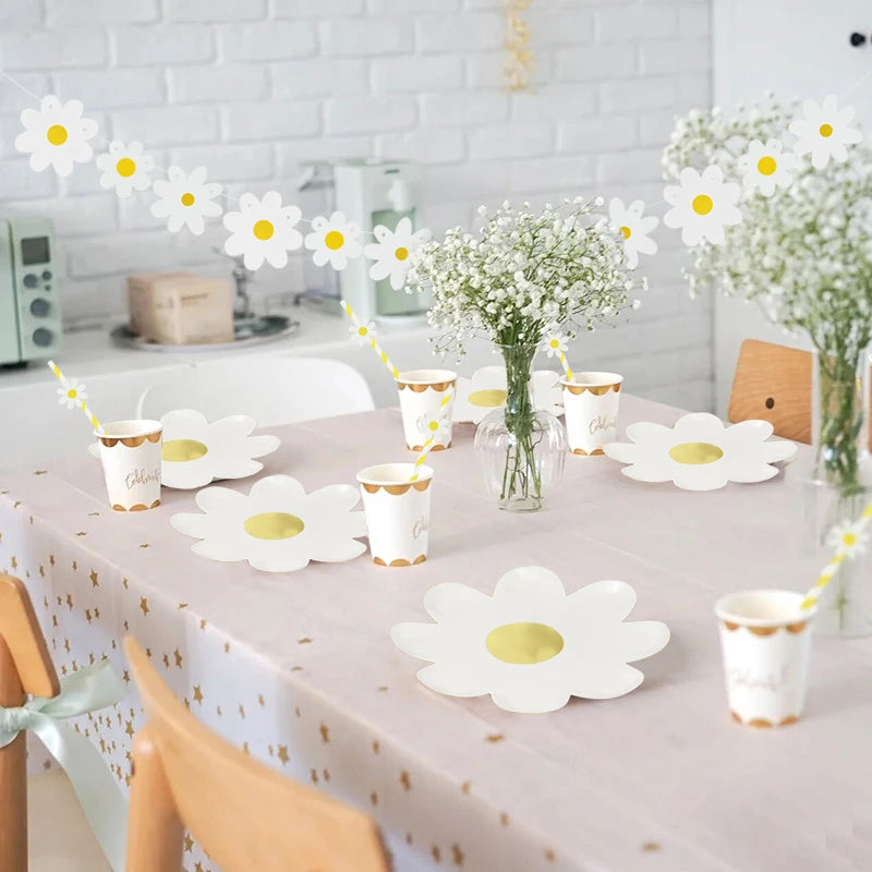 A table set for a party featuring white daisy flower plates, cups, and a floral banner on a pink tablecloth.