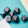 A collection of seven metal polyhedral dice with a dragon font scattered on a teal surface.