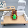 An adorable brown capybara keychain charm with a green accessory resting on a wooden shelf next to a white electronic device.