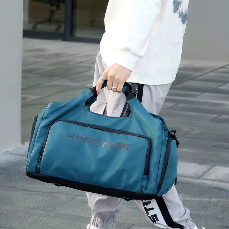 A teal duffel bag with a shoe pocket is carried by a person wearing a white sweatshirt and light pants.