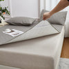 A hand is folding a gray waterproof mattress protector with elastic straps on a bed, next to a pillow.