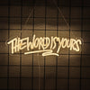 A glowing neon sign spelling out THE WORLD IS YOURS hanging against a dark grid background.