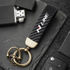 A close-up of a black carbon fiber style leather keychain with a gold swivel buckle resting on a dark slate surface.