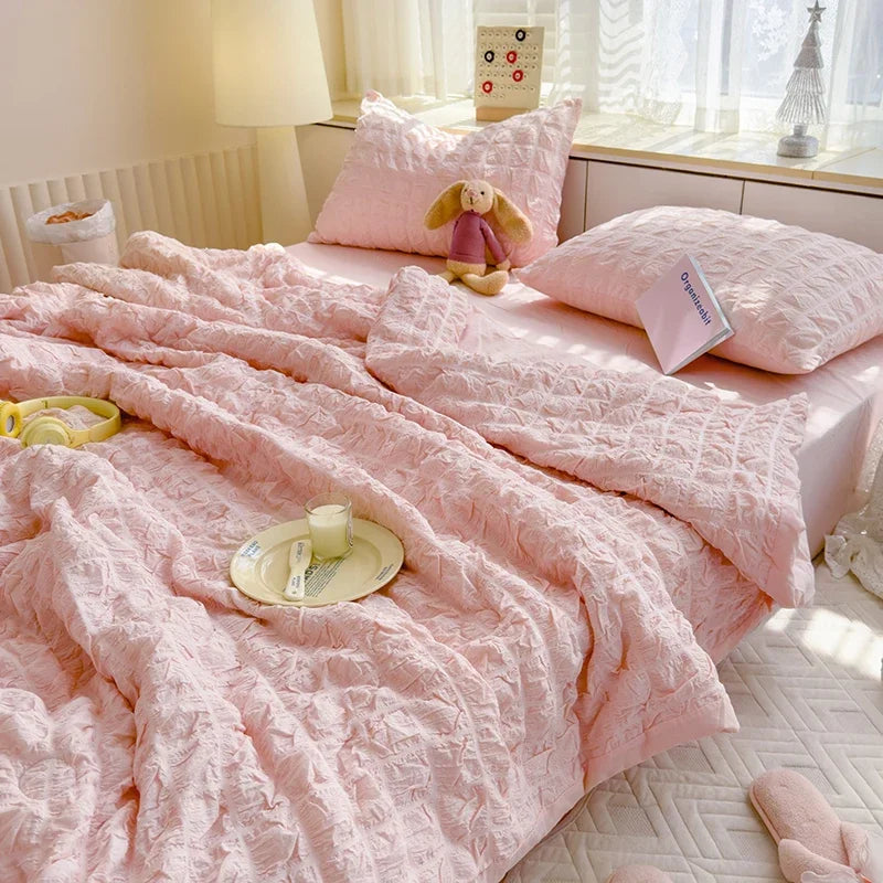 A pink breathable summer seersucker quilt spread on a bed with a stuffed animal and white bedding.