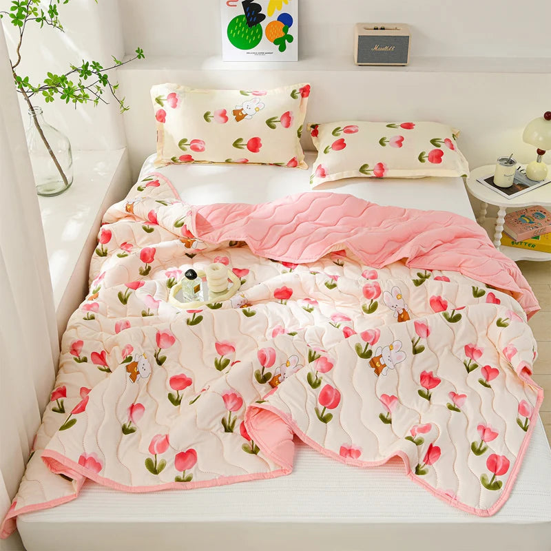 A pink and white quilted summer blanket with a floral pattern is displayed on a white surface next to pillows.