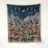 A dark blue woven throw blanket featuring a celestial scene with crescent moons and a field of colorful wildflowers.