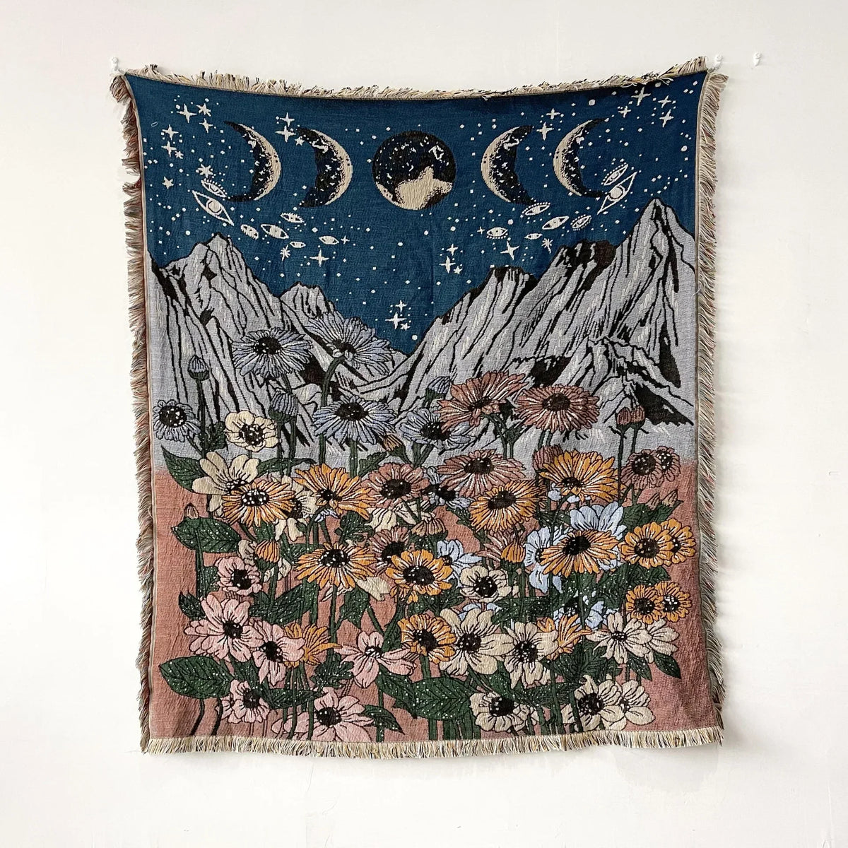 A dark blue woven throw blanket featuring a celestial scene with crescent moons and a field of colorful wildflowers.
