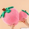 A pink strawberry plush throw pillow with green leaves and a brown stem being held by hands.